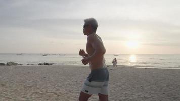 fuerte musculoso anciano asiático trotando a lo largo de la hermosa playa de la isla con gente en el fondo, ambiente de olas y puesta de sol, estilo de vida activo para la mediana edad, seguro de salud, vitalidad de bienestar video