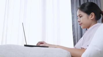 jonge aziatische vrouw freelancer die werkt met een laptopcomputer die de sfeer van het werk thuis laat zien, ga zitten op de bank in de woonkamer op afstand werken vanuit het thuiskantoor, internetverbinding die klanten beantwoordt, browsen op de website video