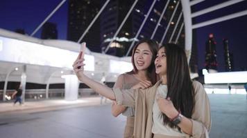 Two attractive asian female tourist making video calling to other friend showing a beautiful modern cityscape behind, distant communication, wireless internet connection and people, holiday vacation