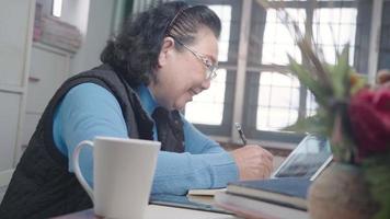 una anciana asiática usa anteojos trabajando desde casa con una lección de clase de cocina en línea de tableta, siéntate a escribir con lápiz en un cuaderno, escena de invierno frío, más delgada distanciada, feliz actividad de ama de casa de jubilación