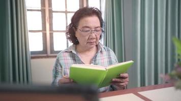 Old asian woman wear glasses reads book at home loving room desk, focusing concentrate while reading, old age learning, reading stories relaxation, middle age wisdom knowledge, free time hobbies video