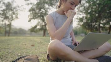 Attraktive Studentin, die allein mit einem Computer-Laptop arbeitet, der auf einer grünen Wiese im Park sitzt, das Lebensziel erreicht, online mit digitaler drahtloser Internettechnologie lernt, natürliches Licht