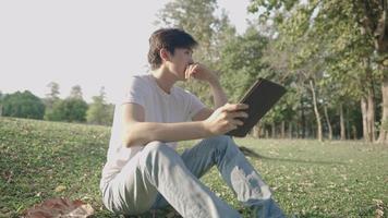jeune homme asiatique à la peau claire utilisant une tablette sans fil assis dans un parc extérieur, par beau temps chaud et ensoleillé, style de vie moderne, application de rencontre en ligne, gadget portable, tendance commerciale