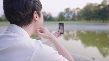 joven feliz sosteniendo un teléfono inteligente, mirando la pantalla, hablando en una videollamada mientras se sienta en el césped del parque, tecnología en línea, aplicación móvil, distanciamiento social, aislamiento video