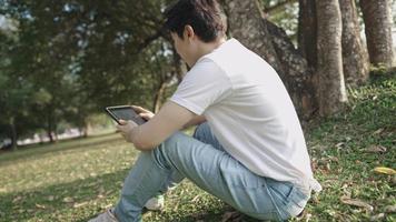 Young asian man in casual clothing sitting with knees up on green meadow inside natural public park, sit under tree shadow during a hot summer, watching online entertainment outdoor, portable device video