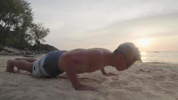 Strong middle aged tan skin man doing bodyweight pushups on the seashore beach, remote workout on the island, silver hair elder active man, outdoor exercises self motivation, morning exercise energy video