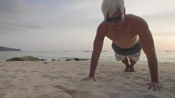 sterke gespierde senior tan huid man doet lichaamsgewicht push-ups op het strand tijdens prachtige zonsondergang, externe training op het eiland, wit haar man maar wordt nooit oud, buiten oefeningen zelfmotivatie video