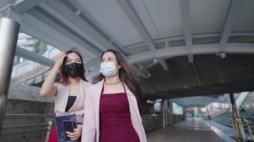 Two young asian working women wear facemask walking along the city crossing bridge urban overpass going to work during rush hour, office employee rush in to work, new normal lifestyle, self protection