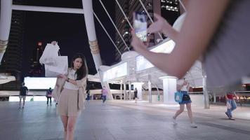 duas amigas asiáticas tirando fotos uma da outra em uma ponte pedonal da cidade moderna durante a noite, dia de compras, venda à meia-noite, crédito e promoção, pessoas e tecnologia de conveniência video
