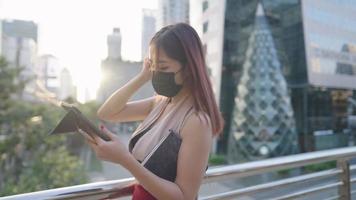 una joven trabajadora asiática que sostiene una tableta digital mira su trabajo inacabado o se conecta en línea de pie en el paso elevado dentro del centro de la ciudad, usuaria de la aplicación, destello de lente solar, mercado de valores video