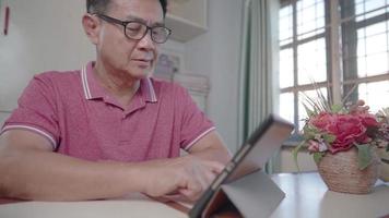hombre asiático de mediana edad con anteojos usando tableta, sentado en la mesa de trabajo en casa dentro de la sala de estar multigeneracional y tecnología, aplicaciones de suscripción, dispositivo portátil de tableta inalámbrica video