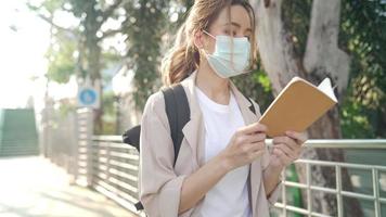 Close up young mixed race woman wearing protective face mask reading while walking in street, beautiful blond hair student reviewing before exam in public park next to school, people and education video