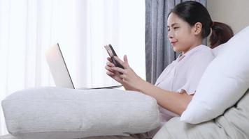 Happy asian young freelance woman smiling using phone and laptop computer while sitting on comfortable bed at home, closeup remote worker uses social media app mobile phone, distant communication tech video