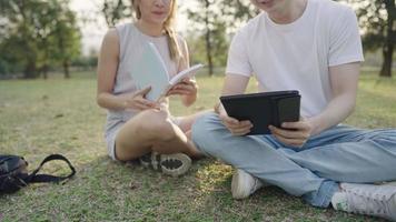 Young colleague working together on the outdoor park, sit down the ground with green grass, discuss brainstorm helping hand on the project, using tablet doing research, classmate reviewing books video