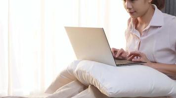 una mujer asiática se sienta de rodillas en una cama cómoda usando una computadora portátil, comienza a trabajar por la mañana, una mujer trabaja en la cama dentro de un dormitorio acogedor, una mujer joven compra en línea y se registra como miembro por un buen trato