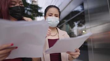 Two asian female office workers discuss works with company paper documents, busy intern and her supervisor, wear face mask while working outside, colleagues best friend relationship, work environment video