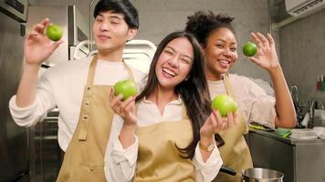 tres jóvenes estudiantes felices en la clase de cocina usan delantales que disfrutan y bromean alegres y divertidos con manzana y naranja en la cocina, sonriendo y riendo, preparando frutas para aprender juntos un curso culinario divertido. video