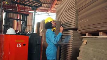 Safety uniform African American Female worker and hard hat inspect storage, factory warehouse stock order, piles of stacking cardboard manufacture, industry product management for logistic transport.