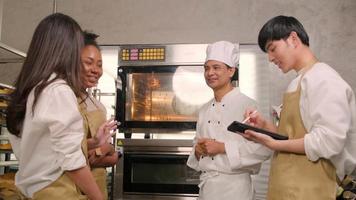El chef asiático senior del curso de aprendizaje culinario enseña y explica sobre el horno eléctrico a los estudiantes de la clase de cocina para hornear masa de pastelería para pan y alimentos de panadería en la cocina de acero inoxidable.