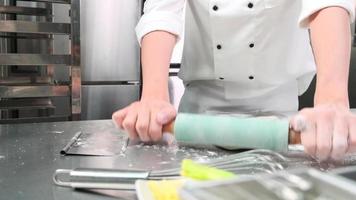 primo piano della mano dello chef in divise bianche da cuoco con grembiuli stanno impastando la pasta con il rullo, preparando pane, torte e cibi freschi da forno, cuocendo in forno nella cucina in acciaio inossidabile del ristorante.
