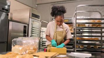entrega de pequenas empresas de inicialização de padaria. uma cozinheira afro-americana está embalando pães e doces feitos à mão e recém-assados em caixas e enviando para compras on-line de clientes na cozinha culinária. video