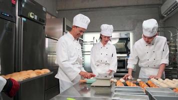 Multiracial professional gourmet team, four chefs in white cook uniforms and aprons knead pastry dough and eggs, prepare bread, and bakery food, baking in oven at stainless steel restaurant kitchen.