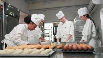 Multiracial professional gourmet team, four chefs in white cook uniforms and aprons knead pastry dough and eggs, prepare bread, and bakery food, baking in oven at stainless steel restaurant kitchen. video