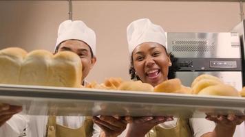 retrato de chefs profissionais de uniforme branco olhando para a câmera com um sorriso alegre e orgulhoso com bandeja de pão na cozinha. um amigo e parceiro de alimentos de panificação e ocupação diária de padaria fresca. video