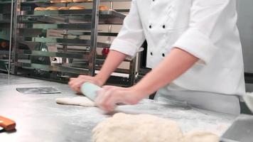 close-up van de hand van de chef in witte kookuniformen met schorten kneden deeg met roller, bereiden brood, taarten en vers bakkerijvoedsel, bakken in de oven in de roestvrijstalen keuken van het restaurant. video