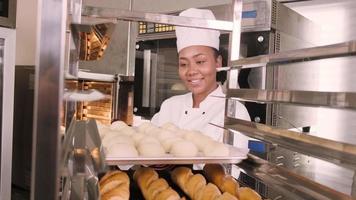 Professionelle afroamerikanische Köchin in weißer Kochuniform, Handschuhen und Schürze, die Brot aus Gebäckteig zubereitet, frische Backwaren zubereitet, im Ofen in der Edelstahlküche des Restaurants backt. video