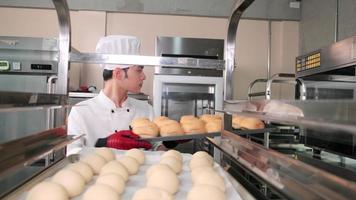 joven chef asiático profesional con uniforme de cocinero blanco con sombrero, guantes y delantal haciendo pan con masa de pastelería, preparando comida fresca de panadería, horneando en el horno en la cocina de acero inoxidable del restaurante. video