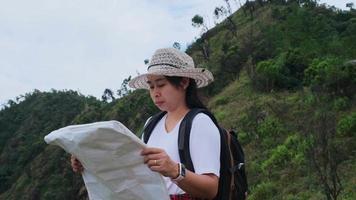Hipster woman with backpack looking at a map on the background of mountains. Female hiker resting in nature and reading a map.