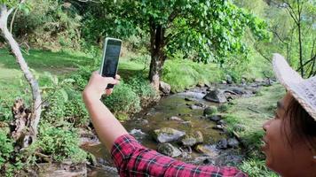 Beautiful Asian woman sits on a wooden bridge across a stream in a mountain forest and makes a video call with a smartphone.