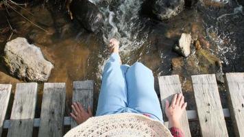 piernas de mujer en jeans sentadas colgando piernas y disfrutando de la naturaleza en un puente de madera sobre un arroyo en un bosque de montaña. vista cercana de piernas balanceándose en un puente de madera. video