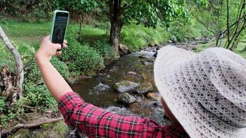linda mulher asiática senta-se em uma ponte de madeira sobre um riacho em uma floresta de montanha e tira uma selfie com um smartphone. estilo de vida de férias e aventuras. video