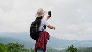 mujer hipster con sombrero relajándose en la naturaleza de vacaciones y tomando selfie con smartphone en el fondo de la montaña.