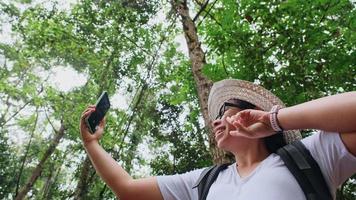 mujer viajera se toma un selfie en un bosque tropical. mujer hipster con mochila usando teléfono móvil en el fondo de frondosos árboles en el bosque. video