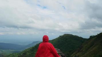 una joven excursionista asiática con un impermeable rojo parada en la cima de la montaña con los brazos levantados y disfrutando de la vista. concepto de viaje y éxito