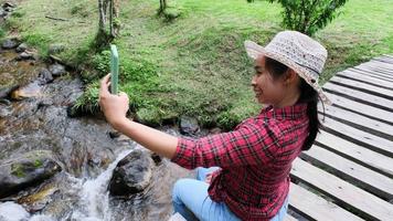 Schöne asiatische Frau sitzt auf einer Holzbrücke über einen Bach in einem Bergwald und macht ein Selfie mit einem Smartphone. Urlaubsleben und Abenteuer. video