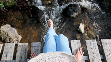 Woman legs in jeans sit hanging legs and enjoying nature on a wooden bridge over a stream in a mountain forest. Close-up view of legs swaying on a wooden bridge. video