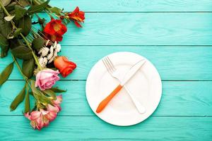 cena festiva con cubiertos, cuchillo, tenedor, plato y rosas sobre fondo de madera azul. vista superior. Bosquejo. flores copie el espacio foto