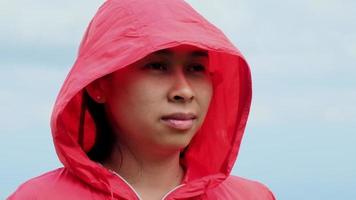 Portrait of a happy Asian female hiker wearing a red waterproof jacket in a hood standing on top of a mountain and and enjoying the view. video
