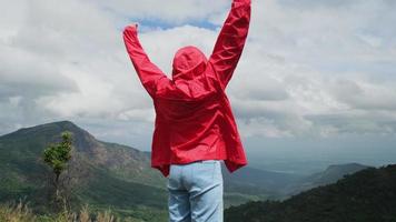 una joven excursionista asiática con un impermeable rojo parada en la cima de la montaña con los brazos levantados y disfrutando de la vista. concepto de viaje y éxito video