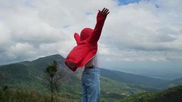 una joven excursionista asiática con un impermeable rojo parada en la cima de la montaña con los brazos levantados y disfrutando de la vista. concepto de viaje y éxito video