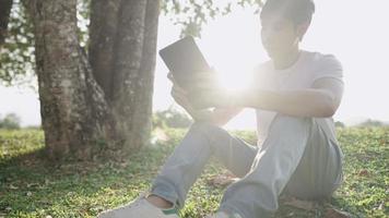 Young asian man sitting under big tree in park with hands holding digital laptop, freelancer in casual clothing remote working outdoor, sit on comfortable meadow with lens flare behind, nature people video