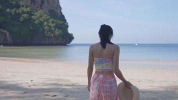 Junge asiatische Frau mit heller Haut, die unter Baumschatten steht, geht hinunter zum Sandstrand, attraktives Mädchen im Urlaub auf der tropischen Insel, Meereslandschaft in Zeitlupe, heißer und sonniger Tag, Reisebuchung video