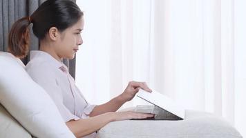mujer trabajadora asiática trabajo cálido con computadora portátil en la cómoda cama y almohadas con luz solar de cortina blanca, mujer trabajando en la cama dentro de un dormitorio acogedor, autora independiente escribiendo en casa video