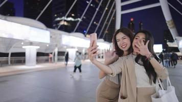 Two cheerful asian tourist waving hand to smartphone with hang up video calling from distant friend, happy fashionable office workers together at famous city bridge in nighttime, wireless internet