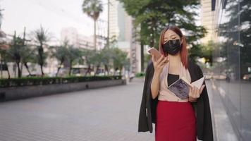 asiatische fröhliche büroangestellte tragen eine medizinische maske, die über kundenanforderungen nachdenkt, telefoniert, eine sekretärin schreibt auf ein papierbuch, eine schöne multitasking-modedesignerin