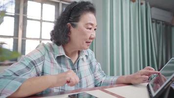 heureuse femme retraitée asiatique moderne utilisant une tablette sans fil, lisant des articles de contenu en ligne, un moment de détente à la maison, un appareil portable, une connexion numérique sans fil, une vieille femme s'adapte à la techno numérique moderne video